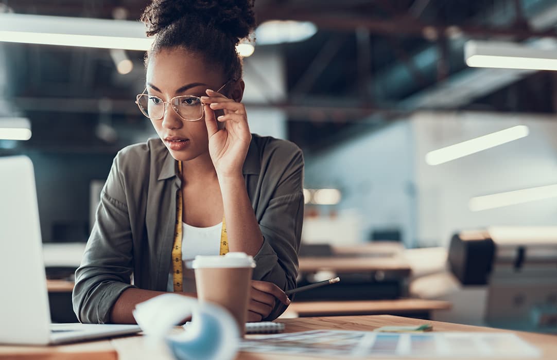 Young black woman with glasses is sitting at her desk at work and looking at a computer