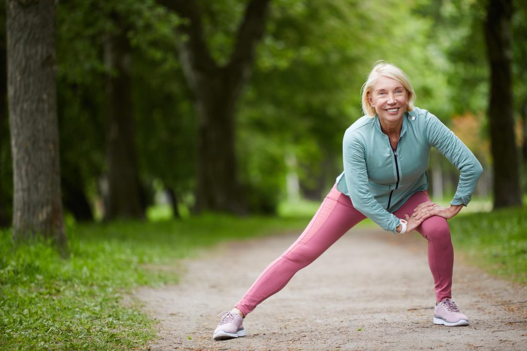Mature cheerful woman in activewear stretching legs while doing physical exercises in a city park among green trees