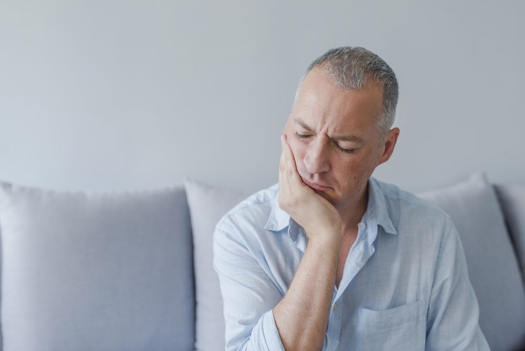 Frustrated young man touching his cheek and keeping eyes closed while sitting on the couch at home